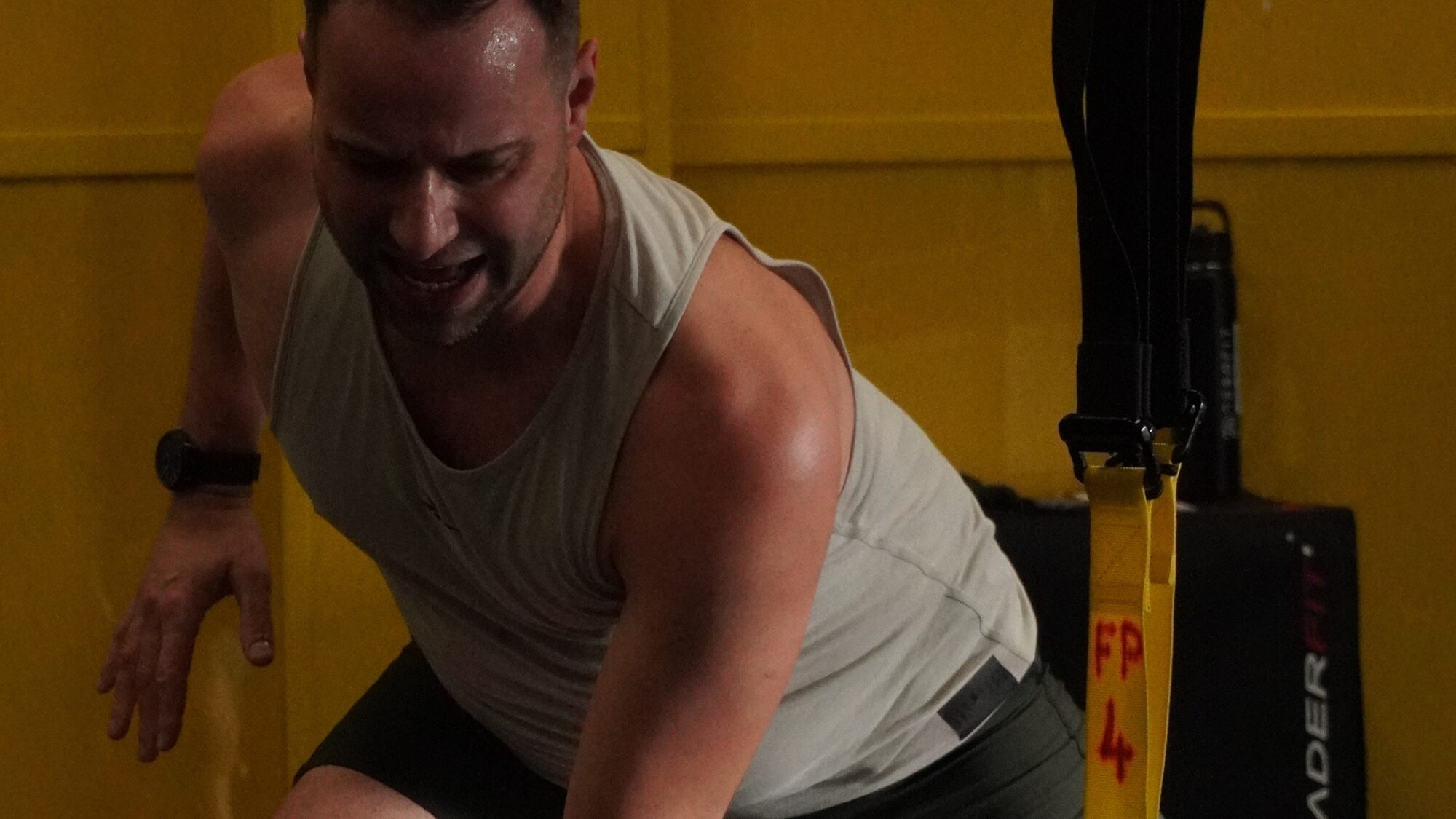 Photo showing Online Personal Trainer David Todd from DTF Virtual Personal Training wearing a grey sleeveless vest with a yellow wall behind and a TRX Suspension trainer in the foreground